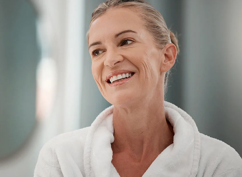 A woman in a white robe smiling and looking relaxed, with a soft focus on the background - Anchored Dentures in Boca Raton, FL