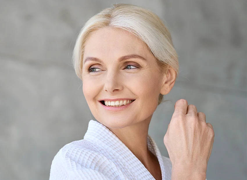 A senior woman with blonde hair smiling and looking away while wearing a white robe - Emergency Dental Procedures in Boca Raton, FL